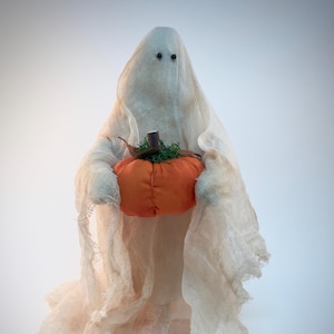 May include: A decorative ghost figurine draped in off-white, semi-sheer cheesecloth fabric. The ghost features two small, dark, circular eyes and holds a small, round, orange fabric pumpkin with a brown stem and green mossy accents.