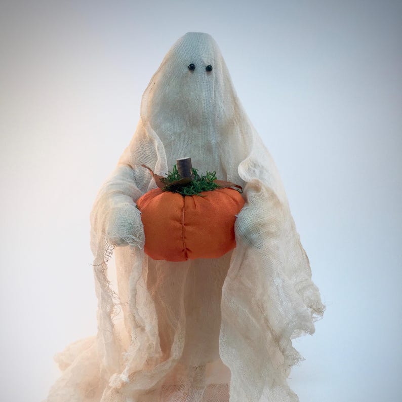 May include: A decorative ghost figurine draped in off-white, semi-sheer cheesecloth fabric. The ghost features two small, dark, circular eyes and holds a small, round, orange fabric pumpkin with a brown stem and green mossy accents.