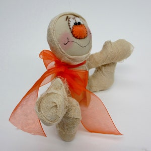May include: A handmade mummy doll with a white fabric body and orange fabric bow. The doll has a stitched-on face with brown eyes and an orange nose.
