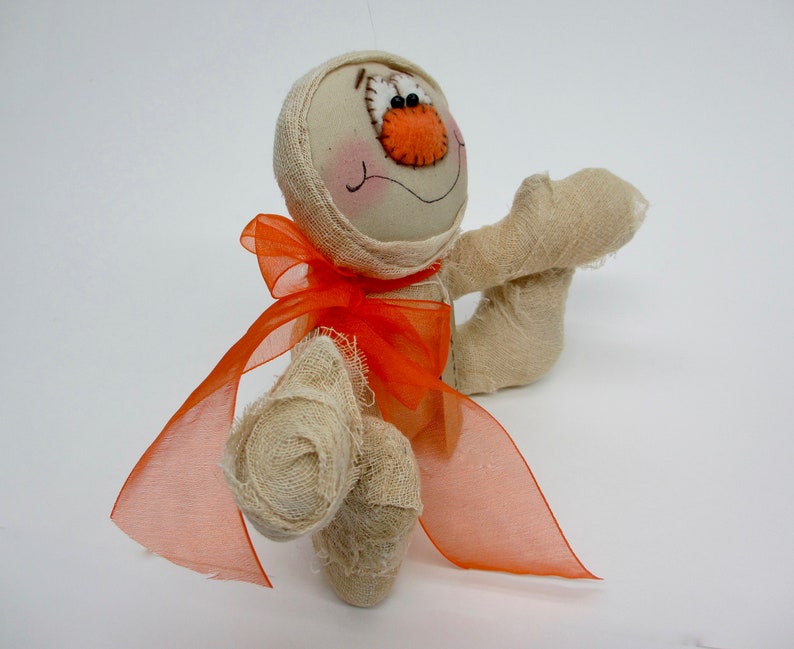 May include: A handmade mummy doll with a white fabric body and orange fabric bow. The doll has a stitched-on face with brown eyes and an orange nose.