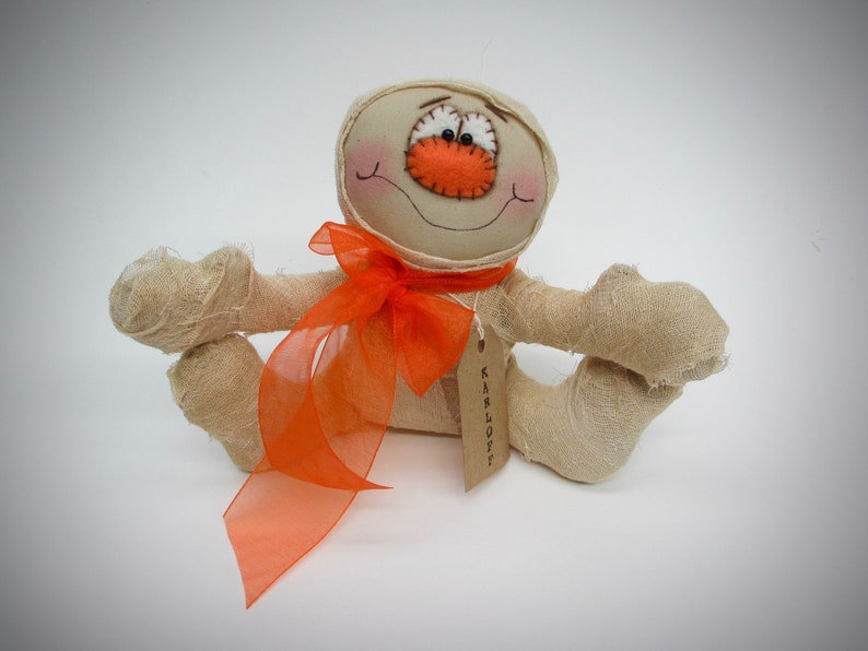 Primitive halloween seated fabric mummy decoration with an orange bow and Karloff tag around his neck