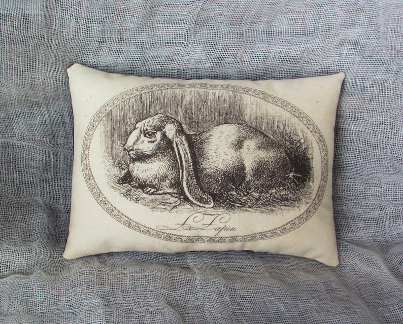 An off white mini pillow with a black illustration of a rabbit with long ears. The illustration is in an oval frame with a decorative border. The text 'L.T. Lapin' is below the illustration.   dimensions are 4" x 6"
