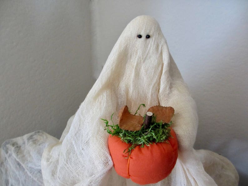 May include: A white fabric ghost figurine with two small black bead eyes. The ghost is draped in a textured, off-white cheesecloth-like material and is holding a small, round, orange fabric pumpkin topped with green moss and brown leaves.
