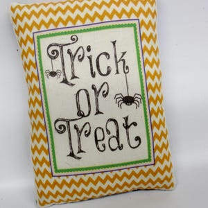 May include: A rectangular decorative pillow featuring a yellow and white chevron pattern border. The center panel is off-white with a green and purple frame, displaying the black text Trick or Treat with two small black spider illustrations.