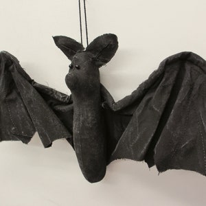 May include: A black fabric bat hanging from a string. The bat has a long body and large wings. The bat has two black eyes.