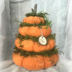 May include: A stacked pumpkin decoration featuring four orange fabric pumpkins of decreasing size, layered with green moss between each tier. A small wooden twig serves as the top stem, and a round white tag with the word Fall is attached.