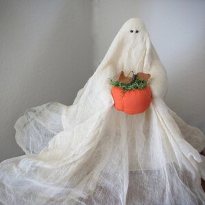 May include: A decorative ghost figurine draped in off-white, sheer, textured fabric. The ghost has two small, dark, circular eyes and holds a bright orange fabric pumpkin topped with green faux moss and small brown felt leaves in its center.