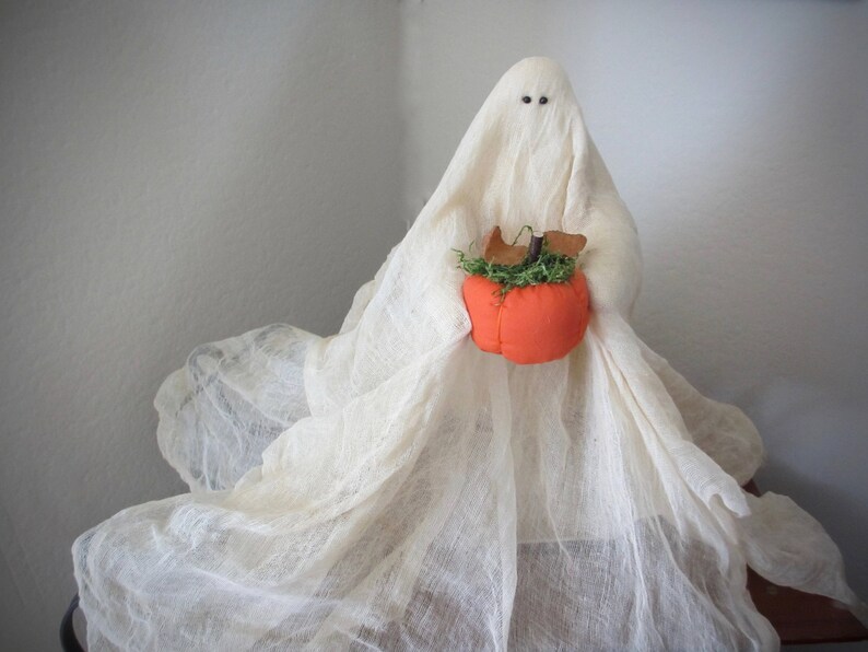 May include: A decorative ghost figurine draped in off-white, sheer, textured fabric. The ghost has two small, dark, circular eyes and holds a bright orange fabric pumpkin topped with green faux moss and small brown felt leaves in its center.