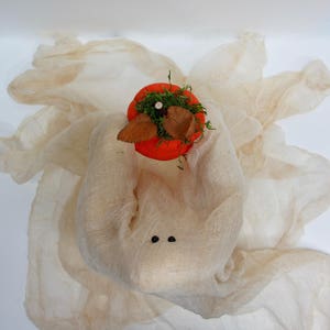 May include: A ghost figure crafted from beige cheesecloth fabric featuring two small black bead eyes. The ghost holds a bright orange pumpkin topper decorated with green moss and two brown dried leaves on its head, creating a seasonal display.
