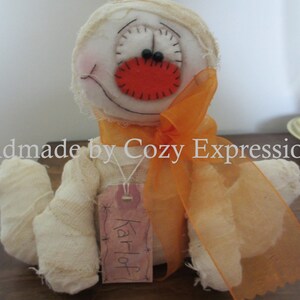May include: A white fabric mummy doll with orange fabric ribbon and a red felt nose. The doll has a tag that says "Karloff".