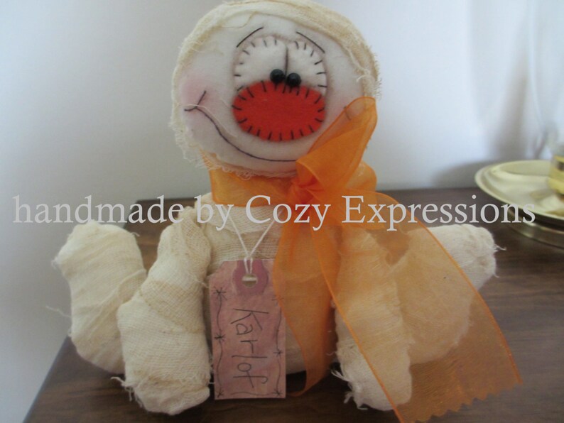 May include: A white fabric mummy doll with orange fabric ribbon and a red felt nose. The doll has a tag that says "Karloff".