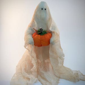 May include: A ghost figurine draped in off-white, semi-sheer fabric with two small black bead eyes. The figure holds a round, orange fabric pumpkin topped with a small brown stem and green moss-like material in its fabric-covered arms.