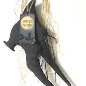May include: A black fabric witch hat ornament featuring a pointed top and a curved brim. The hat is adorned with a rectangular tag displaying a vintage-style illustration of a smiling moon face and dark tree silhouettes, accented by raffia fibers.