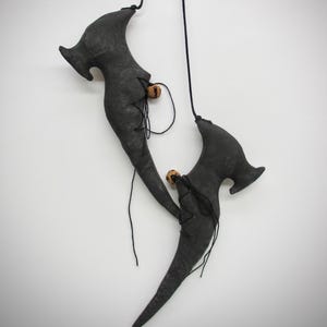 May include: A pair of hanging dark gray fabric shoes with elongated, pointed toes and curved heels. Each shoe features black lace-up detailing and a small, round, light brown wooden bead attached to the laces, suspended by thin black cords.
