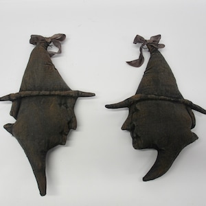 May include: Two dark brown fabric witch hat ornaments with pointed tops and wide brims. Each hat features a tied brown ribbon at the peak and a distressed, textured appearance. The fabric is dark and mottled, giving the items a worn, aged look.