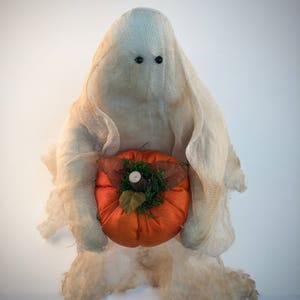 May include: A ghost figurine draped in off-white, textured cheesecloth fabric with two small black bead eyes. The ghost holds a round, bright orange fabric pumpkin topped with green moss, a small brown stem, and two dried brown leaves.