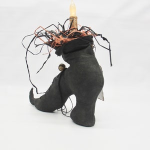 May include: A black witch's boot with a pointed toe and a high heel. The boot is decorated with black and orange fabric and a small, round, gold-colored ornament. A small, white light bulb is attached to the top of the boot.