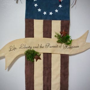 May include: A wooden banner with the text "Life, Liberty and the Pursuit of Happiness" hanging on a brown and white striped fabric background. The fabric has a blue section at the top with white stars.