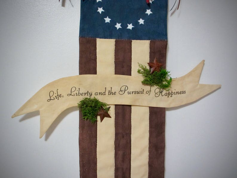 May include: A wooden banner with the text "Life, Liberty and the Pursuit of Happiness" hanging on a brown and white striped fabric background. The fabric has a blue section at the top with white stars.