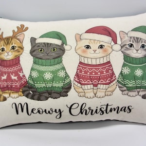 May include: A rectangular throw pillow featuring a printed illustration of four cats wearing holiday sweaters and hats. The cats are adorned in red and green festive attire, with one wearing reindeer antlers. The text Meowy Christmas is printed below.