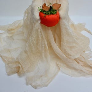 May include: A ghost-shaped figure made of draped, off-white, semi-sheer cheesecloth fabric. The figure holds a small, round, bright orange fabric pumpkin topped with green moss and two brown felt leaves, featuring a dark brown stem at the center.