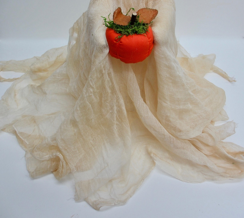 May include: A ghost-shaped figure made of draped, off-white, semi-sheer cheesecloth fabric. The figure holds a small, round, bright orange fabric pumpkin topped with green moss and two brown felt leaves, featuring a dark brown stem at the center.