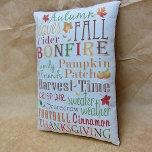 May include: A white pillow with brown trim features a colorful design with the words "Autumn", "Leaves", "Fall", "Cider", "Bonfire", "Family", "Friends", "Pumpkin", "Patch", "Harvest", "Time", "Crisp", "Air", "Sweater", "Scarecrow", "Weather", "Football", "Cinnamon", and "Thanksgiving".