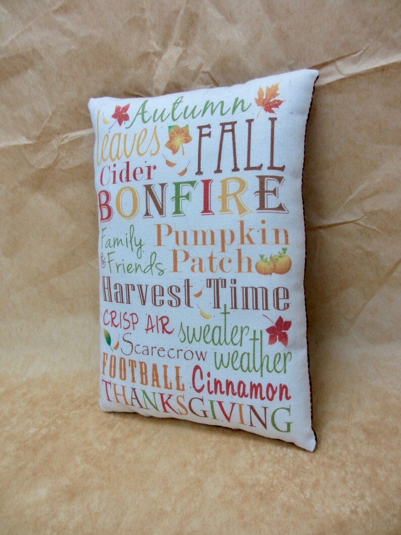 May include: A white pillow with brown trim features a colorful design with the words "Autumn", "Leaves", "Fall", "Cider", "Bonfire", "Family", "Friends", "Pumpkin", "Patch", "Harvest", "Time", "Crisp", "Air", "Sweater", "Scarecrow", "Weather", "Football", "Cinnamon", and "Thanksgiving".