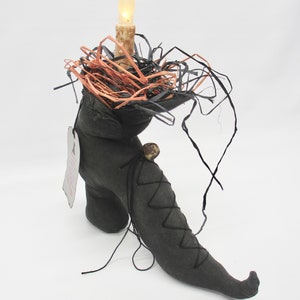 May include: A black witch's boot with a pointed toe and a lit candle on top. The boot is decorated with black and orange straw and a black ribbon.