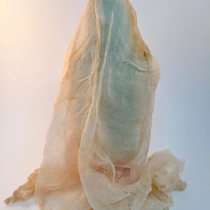 May include: A tall, slender object is completely draped in a sheer, off-white, loosely woven cheesecloth fabric. The fabric hangs in soft, vertical folds, partially obscuring the form beneath while creating a textured, layered appearance.