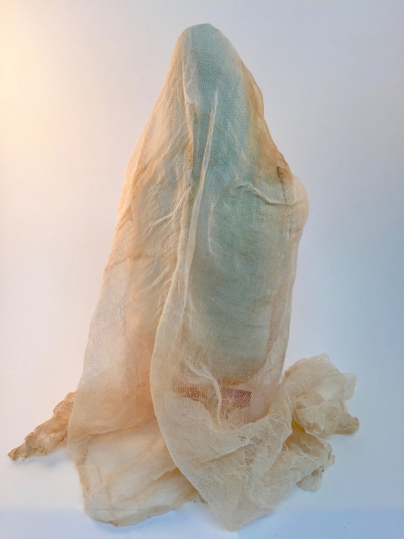 May include: A tall, slender object is completely draped in a sheer, off-white, loosely woven cheesecloth fabric. The fabric hangs in soft, vertical folds, partially obscuring the form beneath while creating a textured, layered appearance.