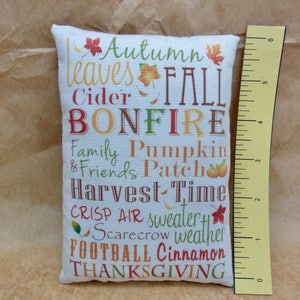 May include: A white pillow with a fall-themed design. The pillow features the words "Autumn leaves Fall Bonfire Family Pumpkin & Friends Patch Harvest Time Crisp Air sweater Scarecrow weather Football Cinnamon Thanksgiving".