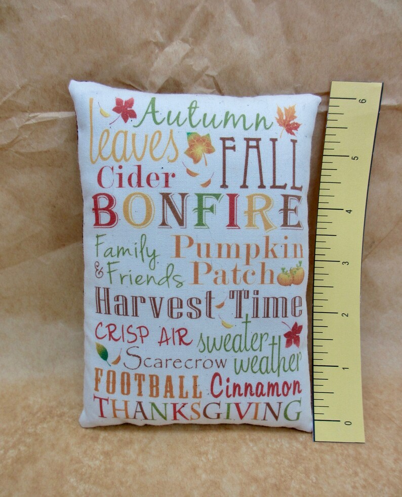 May include: A white pillow with a fall-themed design. The pillow features the words "Autumn leaves Fall Bonfire Family Pumpkin & Friends Patch Harvest Time Crisp Air sweater Scarecrow weather Football Cinnamon Thanksgiving".