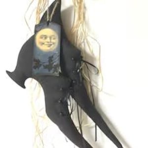 May include: A black fabric ornament shaped like a pointed hat with long, dangling legs, featuring black buttons and lacing details. A rectangular tag hangs from the top, displaying a yellow moon with a face against a dark blue night sky.