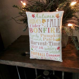 May include: A white pillow with the words "Autumn leaves FALL BONFIRE Family Pumpkin & Friends Patch Harvest Time CRISP AIR sweater weather FOOTBALL Cinnamon THANKSGIVING" printed on it in various colors.