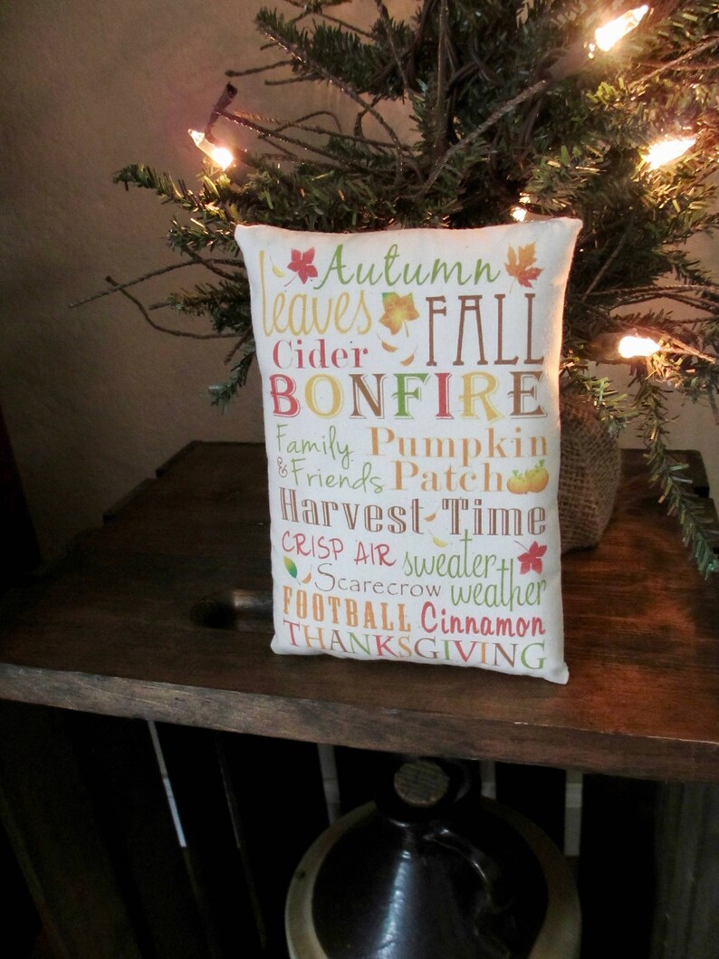 May include: A white pillow with the words "Autumn leaves FALL BONFIRE Family Pumpkin & Friends Patch Harvest Time CRISP AIR sweater weather FOOTBALL Cinnamon THANKSGIVING" printed on it in various colors.