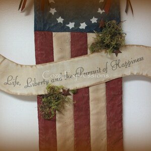May include: A rustic American flag with a banner that reads "Life, Liberty and the Pursuit of Happiness". The flag is made of red, white, and blue fabric and has a distressed look. The banner is made of white fabric and has a brown inscription.