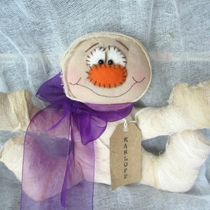 May include: A handmade mummy doll with a purple ribbon and a tag that says "KARLOFF". The doll has a stitched-on orange nose and white eyes.
