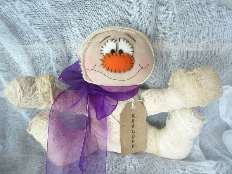 May include: A handmade mummy doll with a purple ribbon and a tag that says "KARLOFF". The doll has a stitched-on orange nose and white eyes.