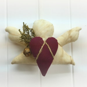 May include: A white fabric angel with outstretched wings holding a red heart with twine wrapped around it. The angel has a sprig of dried greenery and a gold bell attached to its wing.