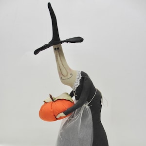May include: A fabric witch doll with a long neck, wearing a black pointed hat and a black dress with a white lace collar and a sheer white apron. The doll holds a stuffed orange pumpkin with a brown stem in its arms against a plain background.