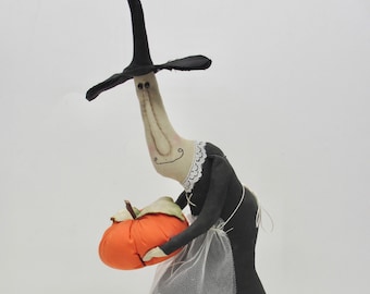 Puede incluir: Una muñeca bruja de tela con cuello largo, que lleva un sombrero puntiagudo negro y un vestido negro con cuello de encaje blanco y un delantal blanco transparente. La muñeca sostiene una calabaza naranja rellena con tallo marrón en sus brazos.