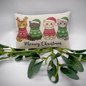 May include: A decorative pillow featuring four cartoon cats dressed in Christmas sweaters and hats. The cats are arranged in a row, with the text "Meowy Christmas" below them. A sprig of mistletoe is in the foreground.