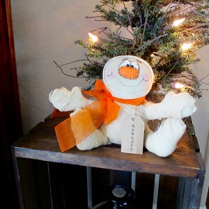 May include: A white fabric mummy doll with orange ribbon and a tag that says "KARLOFF" sitting on a wooden crate in front of a small Christmas tree with white lights.