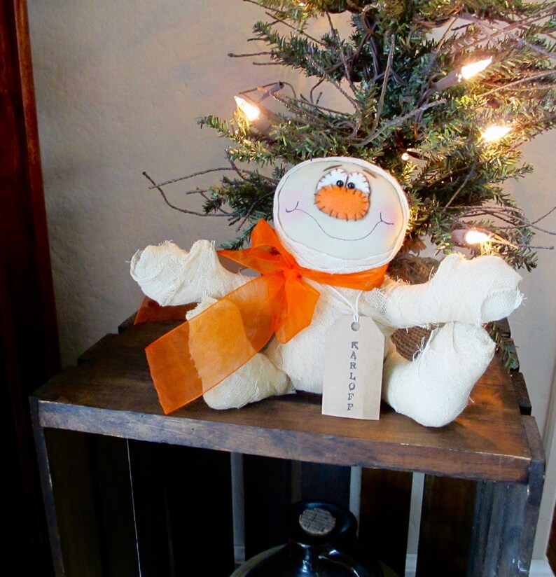 May include: A white fabric mummy doll with orange ribbon and a tag that says "KARLOFF" sitting on a wooden crate in front of a small Christmas tree with white lights.