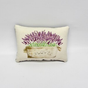Puede incluir: Una almohada blanca con un dise&ntilde;o de flores de lavanda en una maceta con la palabra "FLOWER" impresa.