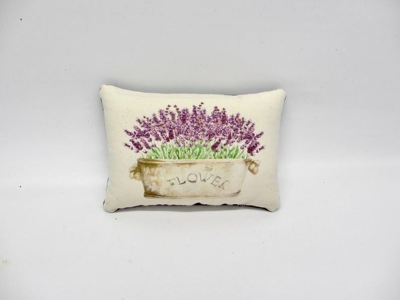 Puede incluir: Una almohada blanca con un dise&ntilde;o de flores de lavanda en una maceta con la palabra "FLOWER" impresa.