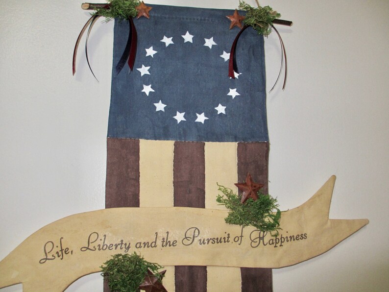 May include: A decorative American flag with 13 stripes and a blue canton featuring 13 white stars in a circle. The flag is hanging from a wooden dowel with a banner below that reads "Life, Liberty and the Pursuit of Happiness".