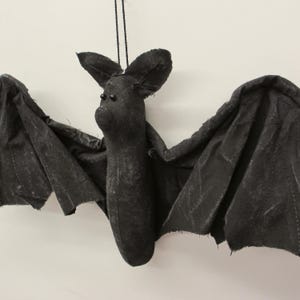 May include: A black fabric bat ornament with a soft, stuffed body and large, pointed ears. The bat features two small, round black bead eyes and wide, outstretched wings made of dark, textured fabric with frayed edges, suspended by a thin black string.