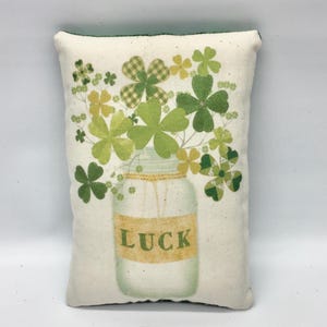 May include: A rectangular off-white fabric pillow featuring a printed illustration of a glass mason jar filled with various green and yellow clover leaves. The jar has a yellow label across the center with the word LUCK printed in green letters.